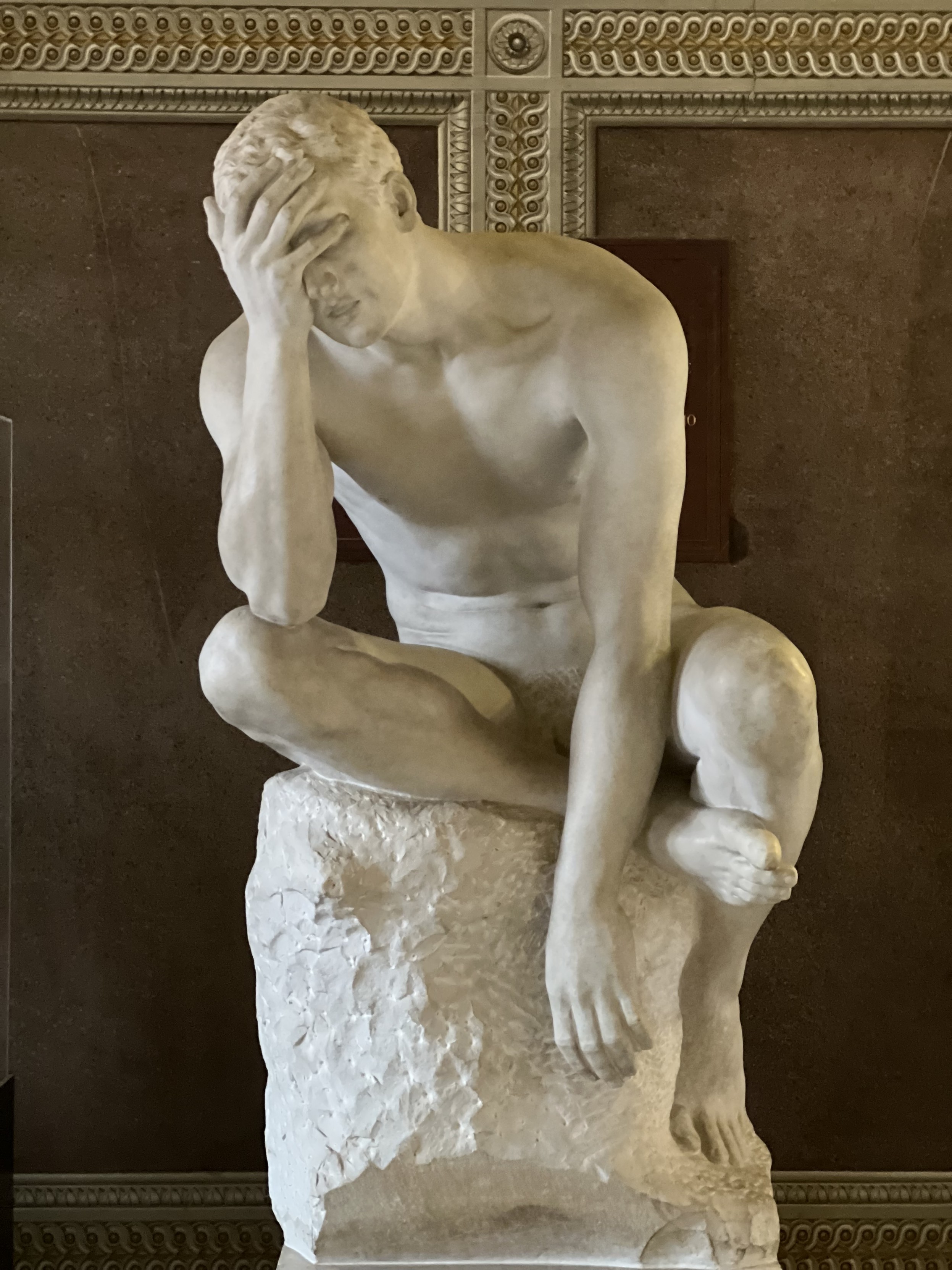 Roman Era sculpture of a man, sitting on a pedestal, burying his downcast face in his right hand.